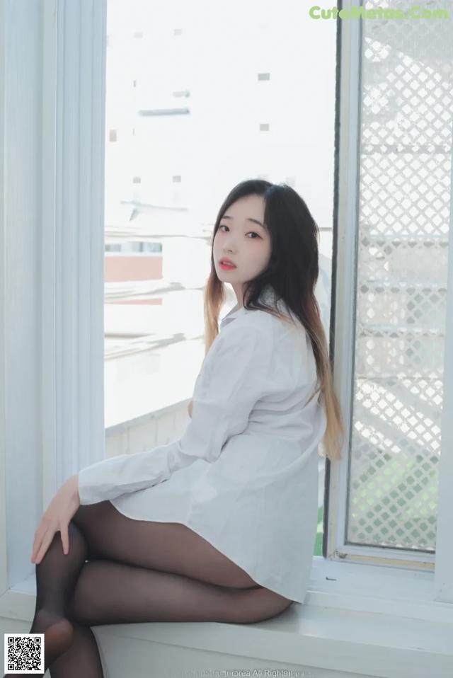 A woman sitting on a window sill in a white shirt and black tights.