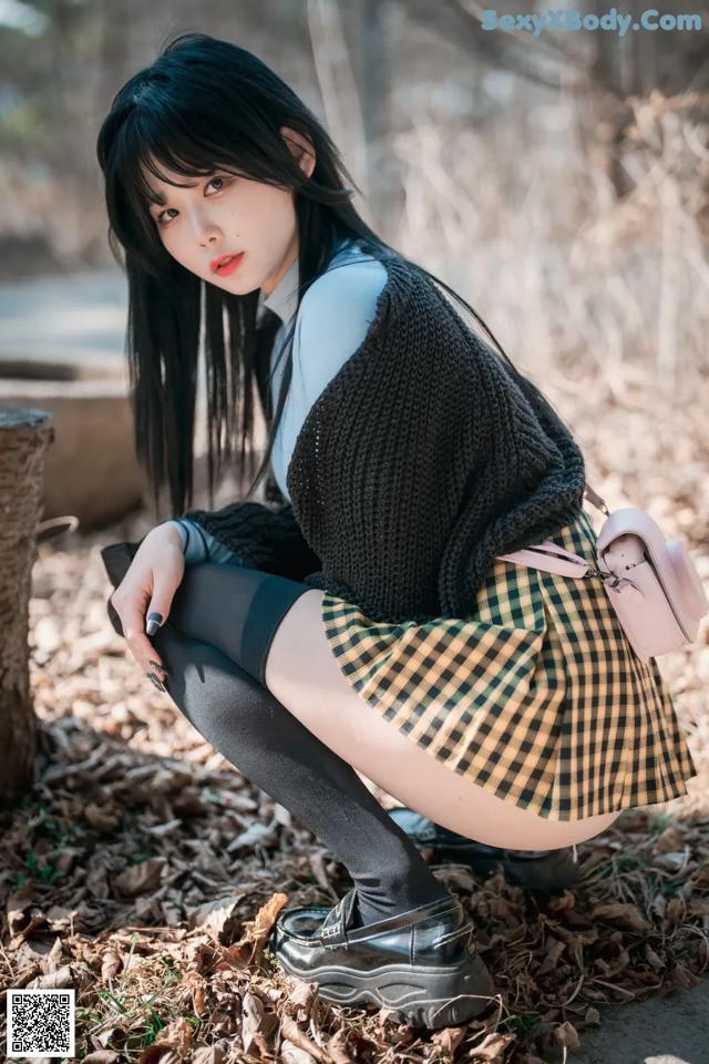 A woman in a plaid skirt crouching down in the woods.